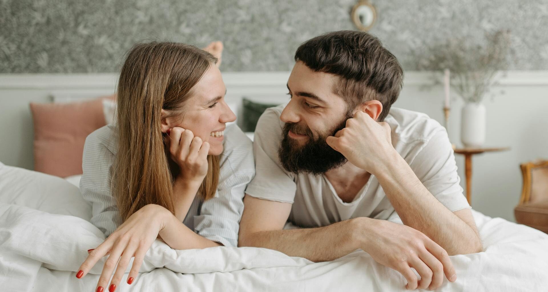 Happy couple lying face to face on a bed, sharing smiles in a cozy and intimate bedroom setting.