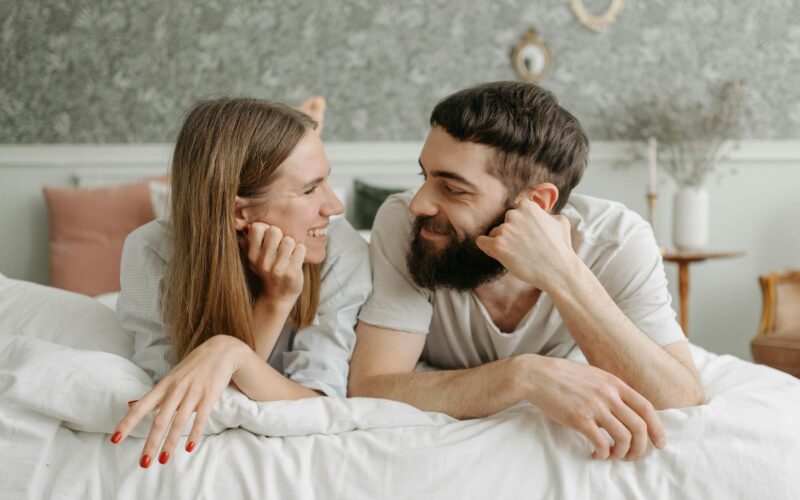 Happy couple lying face to face on a bed, sharing smiles in a cozy and intimate bedroom setting.