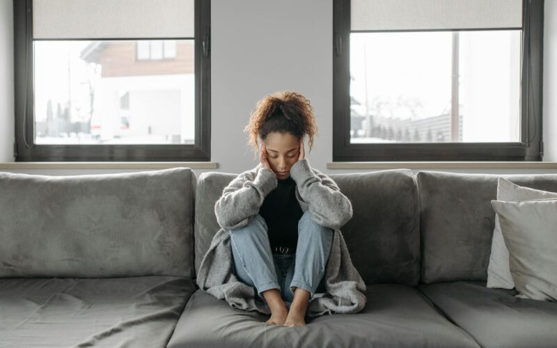 A woman sits on a sofa looking unwell, emphasizing a sense of illness or fatigue indoors