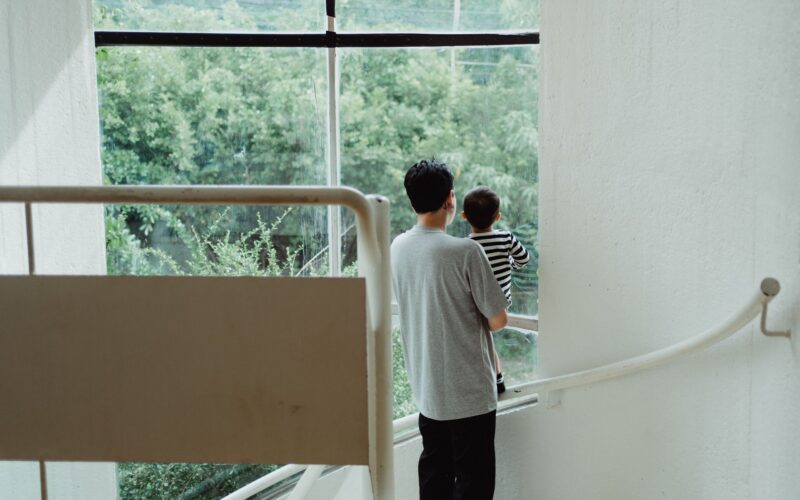 A man holding a child looks out a large window, standing on a staircase.