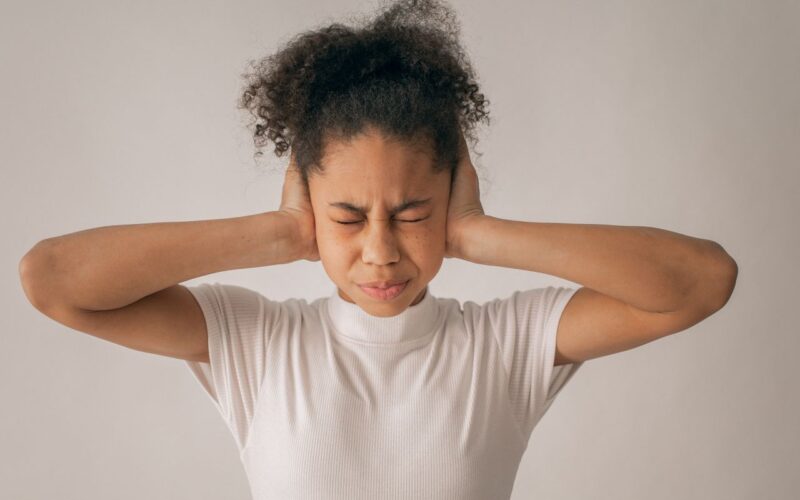 Teen girl with eyes closed and hands on ears in studio, feeling stressed.