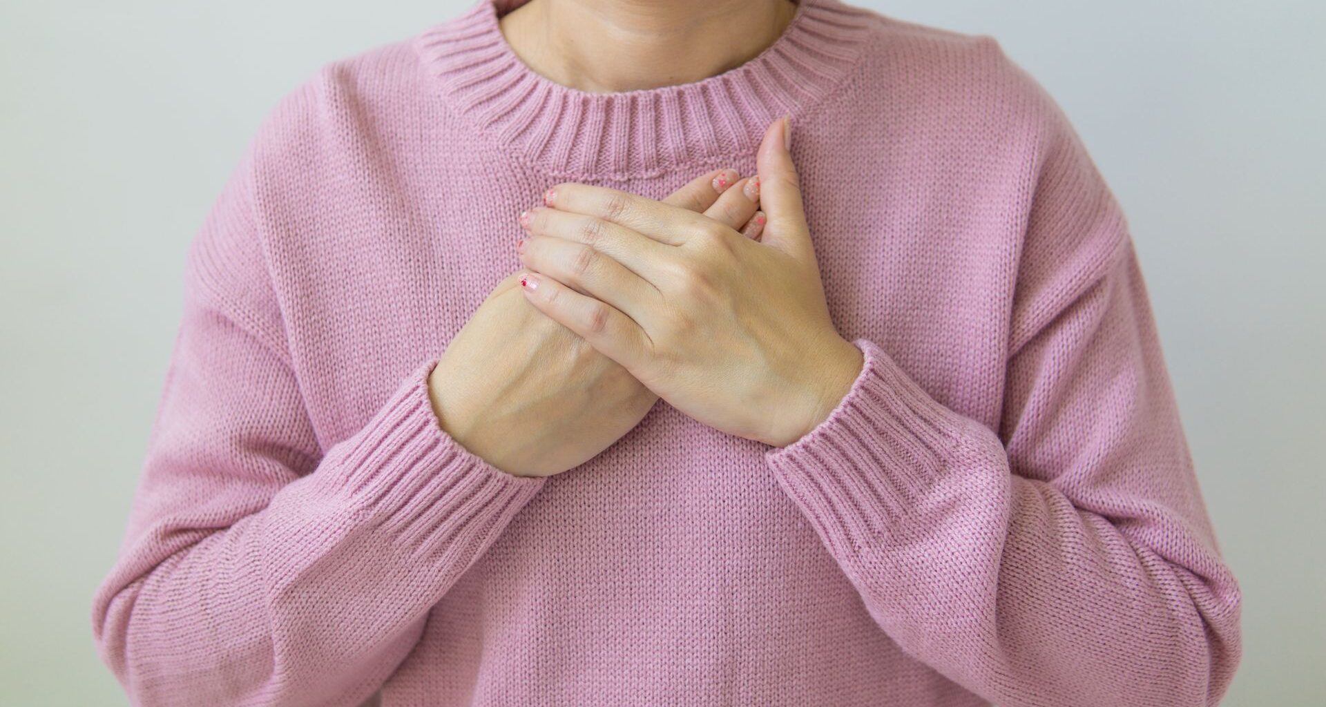 A woman gently places her hands on her chest, wearing a soft pink sweater, symbolizing care and wellness