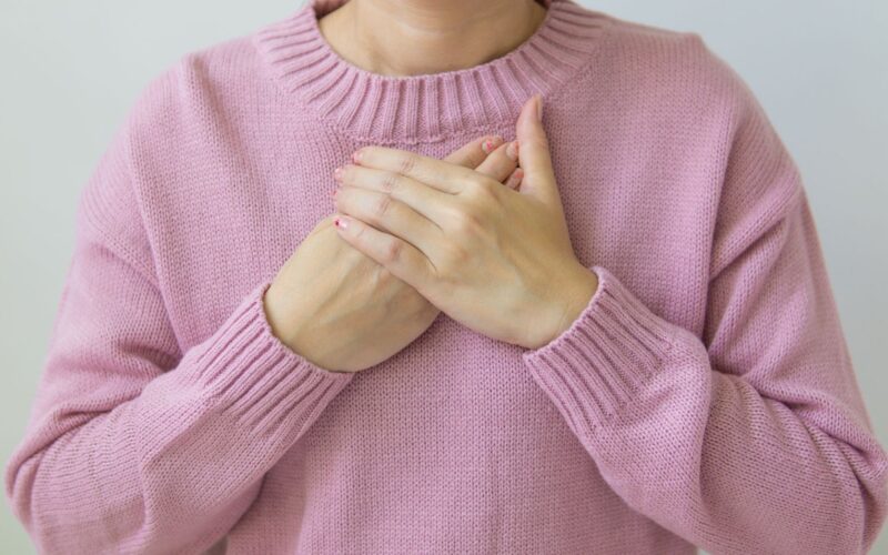 A woman gently places her hands on her chest, wearing a soft pink sweater, symbolizing care and wellness